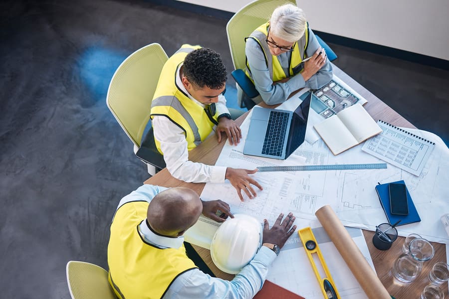 A team of construction professionals in high-visibility vests collaborating over blueprints and a laptop.