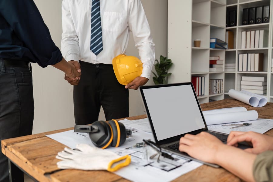 Two construction professionals shaking hands over project documents, with a laptop and protective gear on the desk.